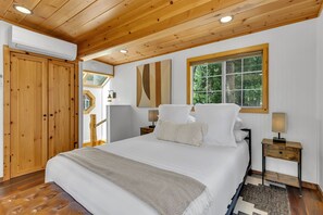 A bedroom with a large bed, wooden ceiling, a window with a view of trees, and a wooden wardrobe. - La Petite - cozy fireplace cabin in town (Idyllwild-Pine Cove)