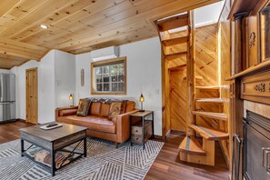 A cozy living room with a wooden ceiling, a leather sofa, a coffee table, and a fireplace with a wooden staircase. - La Petite - cozy fireplace cabin in town (Idyllwild-Pine Cove)