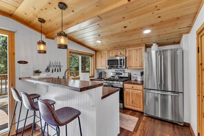 A modern kitchen with wooden cabinets, stainless steel appliances, and a bar area with stools. - La Petite - cozy fireplace cabin in town (Idyllwild-Pine Cove)