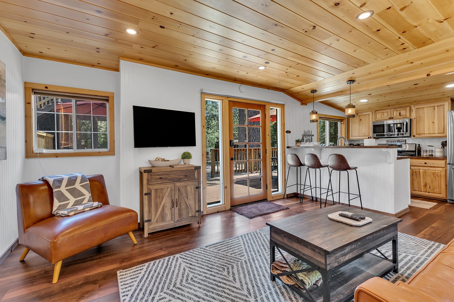 A spacious living area with a wooden ceiling, a leather sofa, a TV mounted on the wall, and a kitchen area with bar stools.
