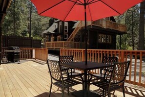 Una terraza de madera con una mesa y sillas, un paraguas rojo y una cabaña al fondo.