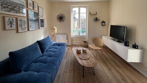 A living room with a blue sofa, a coffee table, a TV on a white cabinet, and a window with a view of buildings.