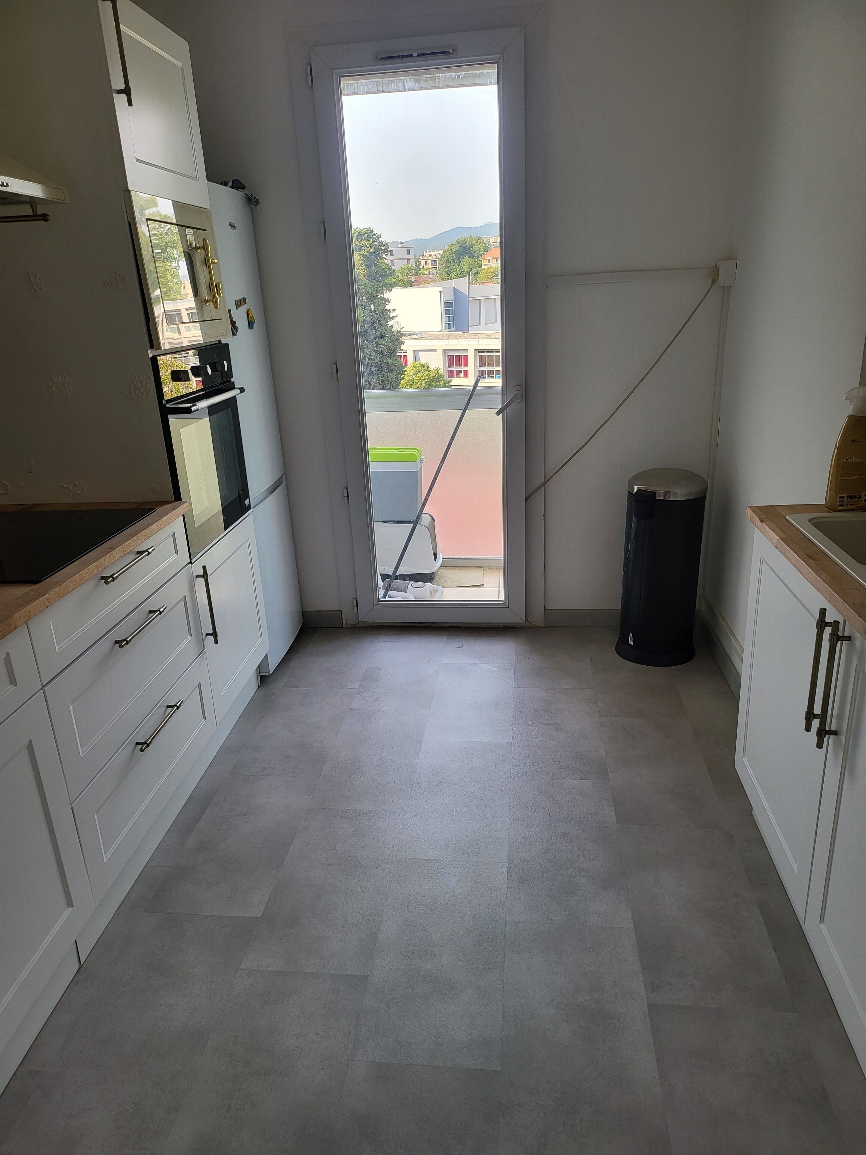 Una cocina con armarios blancos, un bote de basura negro y vistas de edificios a través de una puerta.