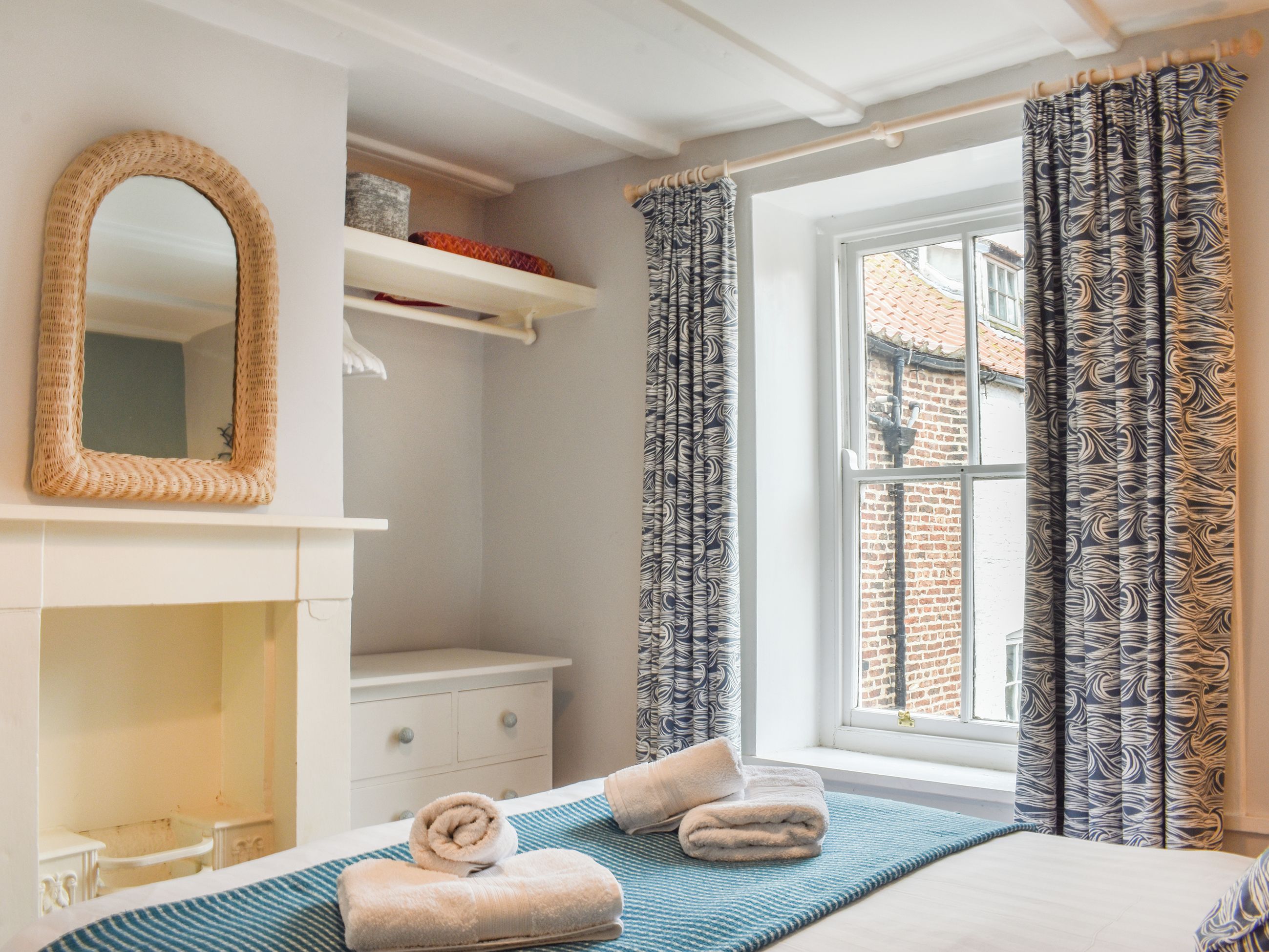 A bedroom with a bed, a mirror, a shelf with decorative items, and a window with curtains.