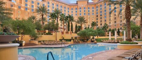 A hotel courtyard with a swimming pool, lounge chairs, palm trees, and a multi-story building.