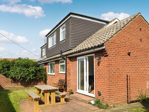 A two-story house with a brick exterior and a grey roof. There is a wooden patio with a table and chairs, and a sliding glass door leading to the interior. - You will love this spacious semi-detached dormer bungalow with enclosed garden. (Flamborough, near Bridlington)