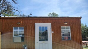 A small wooden building with a white door, two windows, and a small porch area. - The Cricket Cabin Bathroom sep building (Kempner)