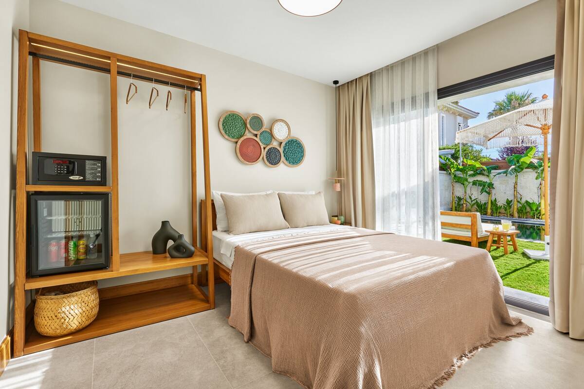 Une chambre avec un lit, un mini-bar, une étagère en bois et une vue sur un jardin, visible à travers une grande fenêtre.