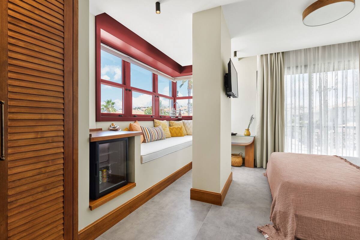 Une chambre d’hôtel dotée d’une grande fenêtre, d’un téléviseur intégré, d’un lit et d’un petit coin salon avec des coussins.