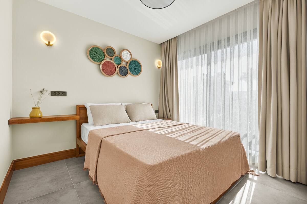 Une chambre moderne avec un grand lit, une table de chevet, des lampes fixées au mur et une fenêtre avec des rideaux.