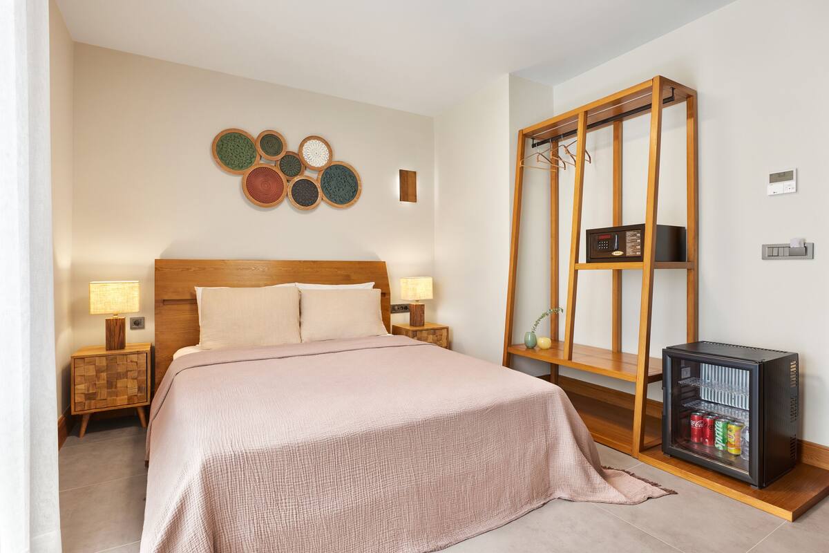 Une chambre bien rangée, avec un lit, des lampes de chevet, une œuvre d’art accrochée au mur, une armoire en bois et un mini-réfrigérateur.