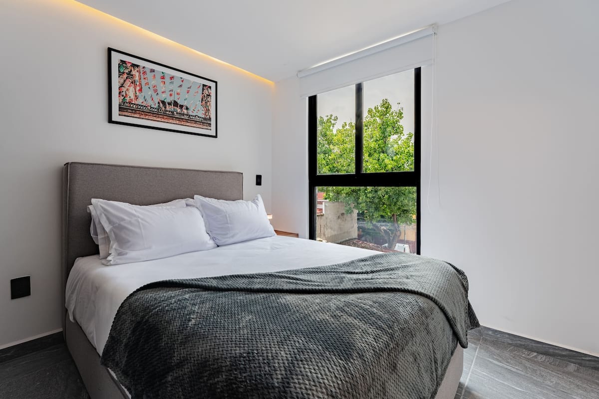 A modern bedroom with a large bed, a framed picture on the wall, and a window with a view of greenery.