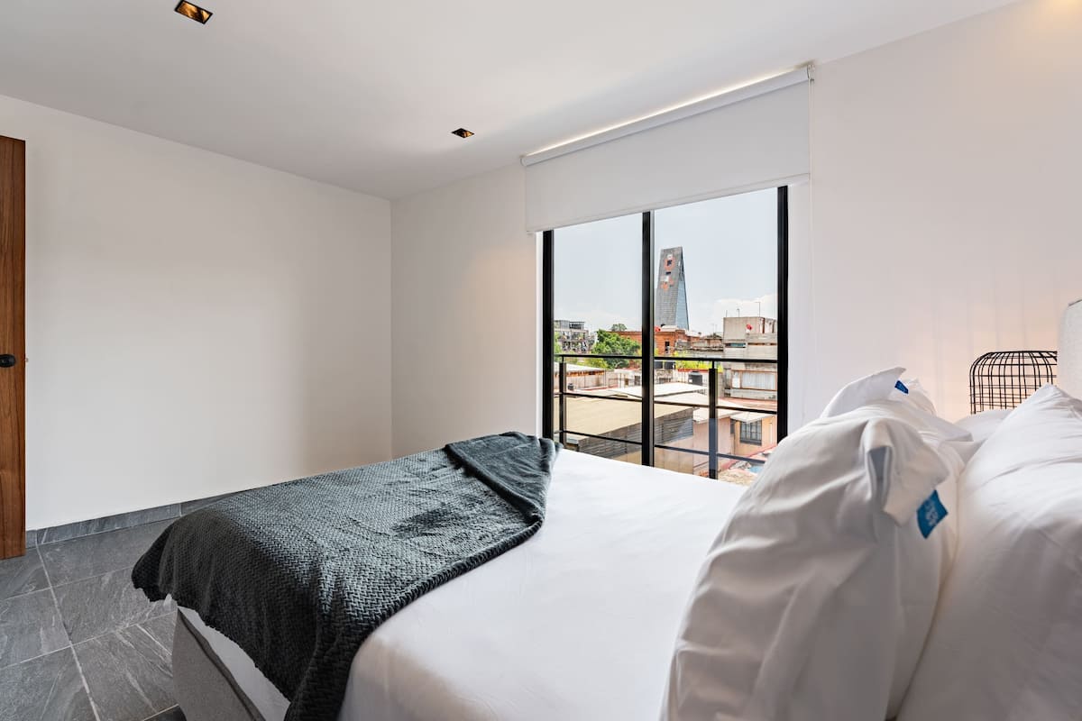 A hotel room with a bed, a window with a view of buildings, and a door.