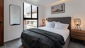 Een moderne slaapkamer met een groot bed, een raam met uitzicht op gebouwen, een nachtkastje met een lamp en een ingelijste foto aan de muur.