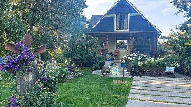 Une maison avec un toit bleu, un porche à l’avant et un jardin où poussent différentes plantes et fleurs.