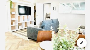 Una sala de estar moderna con un sofá gris, un sillón marrón y un televisor montado en una pared blanca. El suelo es de madera con un diseño en espiga, hay una alfombra blanca y una planta en la esquina.
