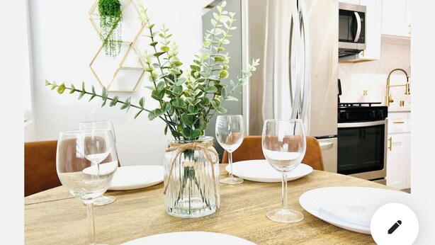 Una mesa de comedor dispuesta con platos blancos y copas de vino, un tarro de cristal con follaje, y una cocina al fondo.