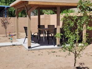 Een overdekt terras met een tafel en stoelen, omgeven door een bakstenen muur en een kleine boom.