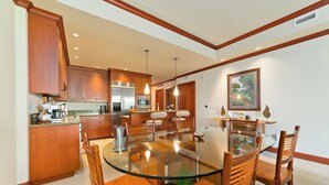 A modern kitchen with wooden cabinets, a glass dining table, and a central island. - Kolea - Luxe 2BR Condo with Pool Access (Waikoloa)