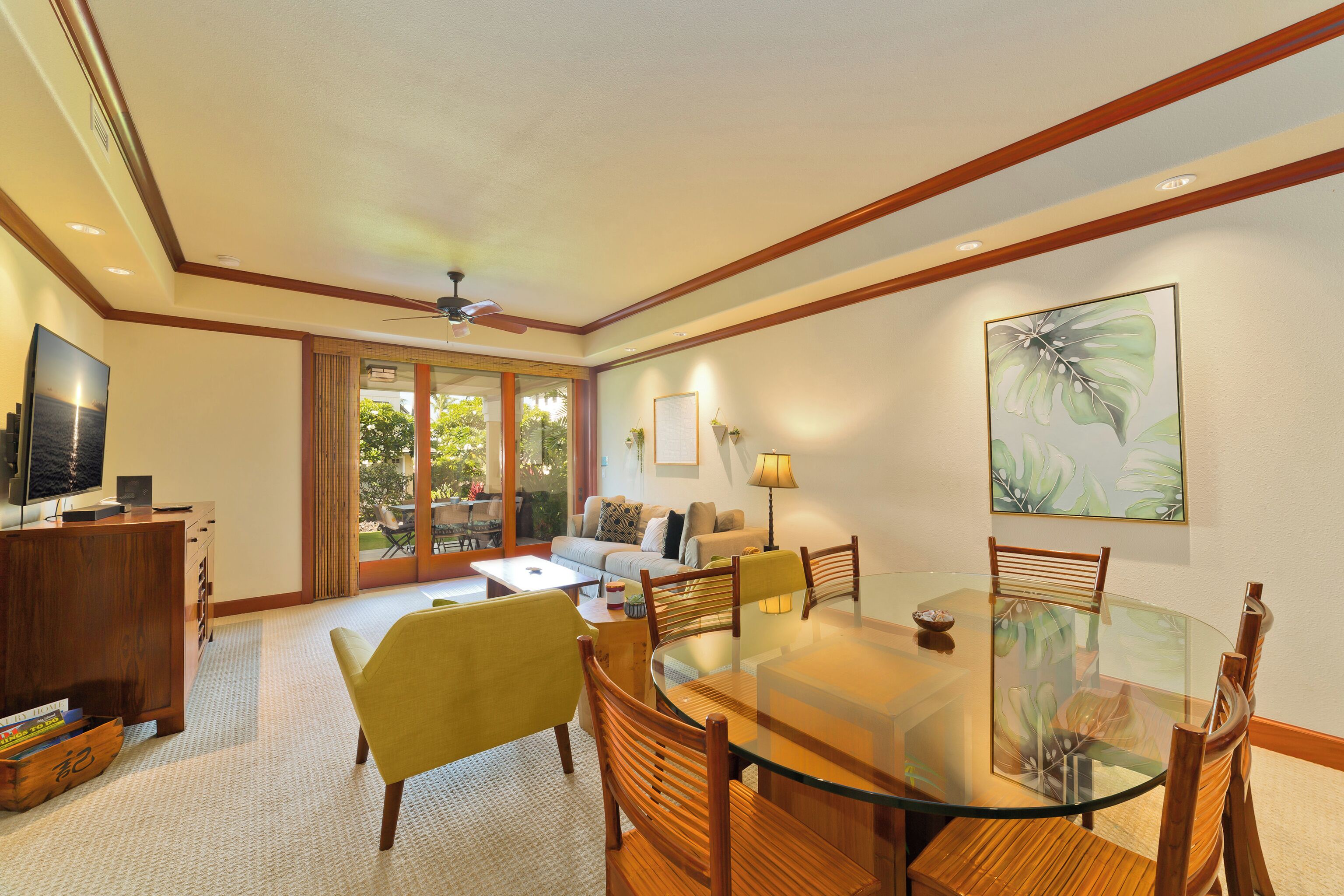 A spacious living area with a glass dining table, wooden chairs, a sofa, and a television on a wooden cabinet.