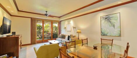 A spacious living area with a glass dining table, wooden chairs, a sofa, and a television on a wooden cabinet.