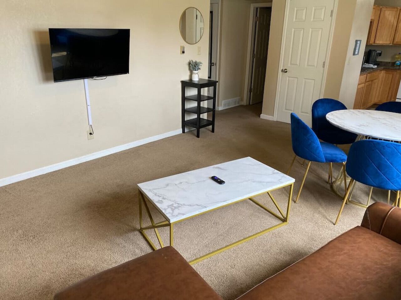 Ein Wohnzimmer mit an der Wand montiertem Fernseher, einem kleinen Tisch mit Pflanze, einem runden Tisch mit blauen Stühlen und einem braunen Sofa.