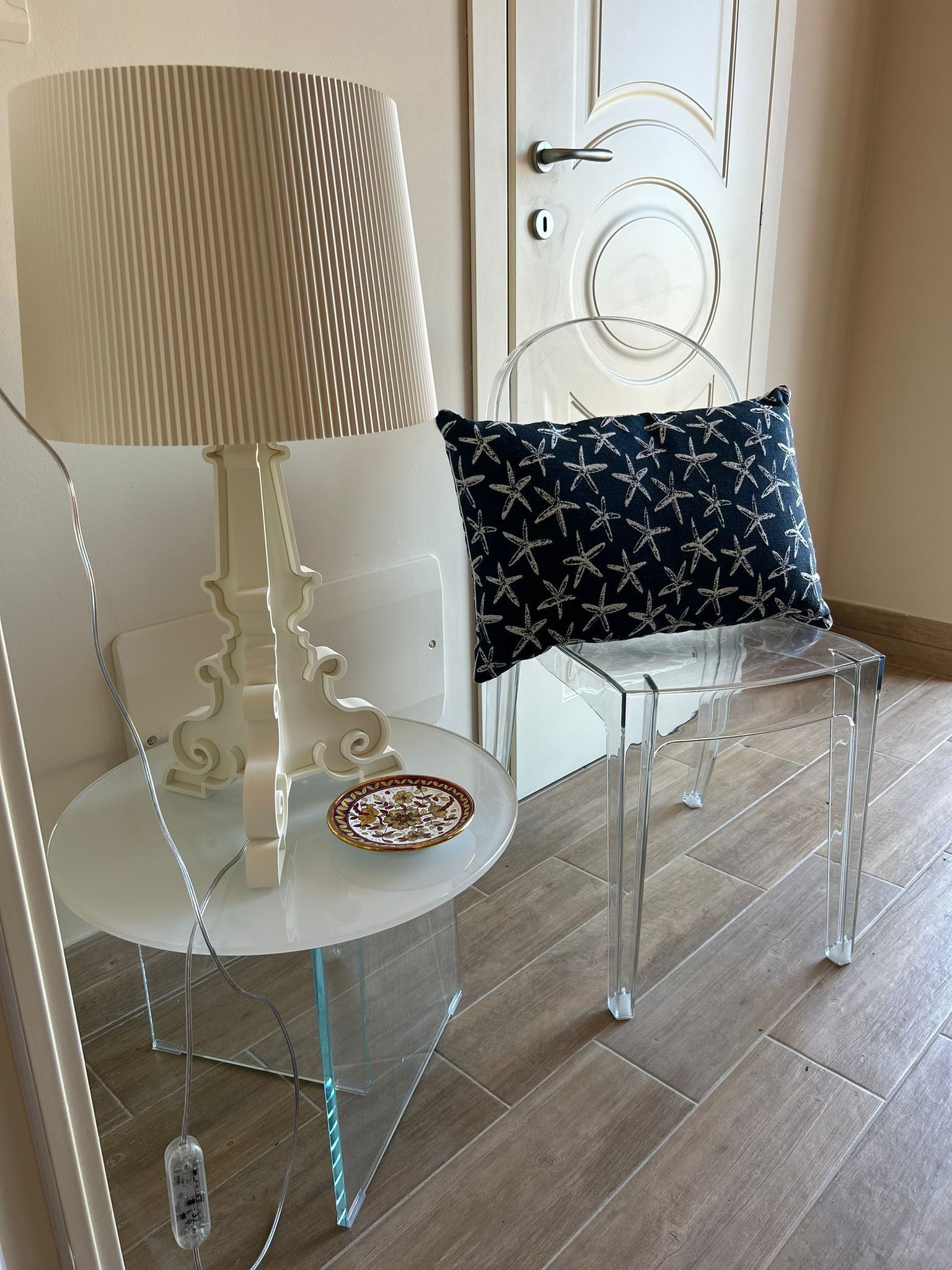A modern room with a clear glass table, a white chair, a black patterned pillow, and a white lamp.