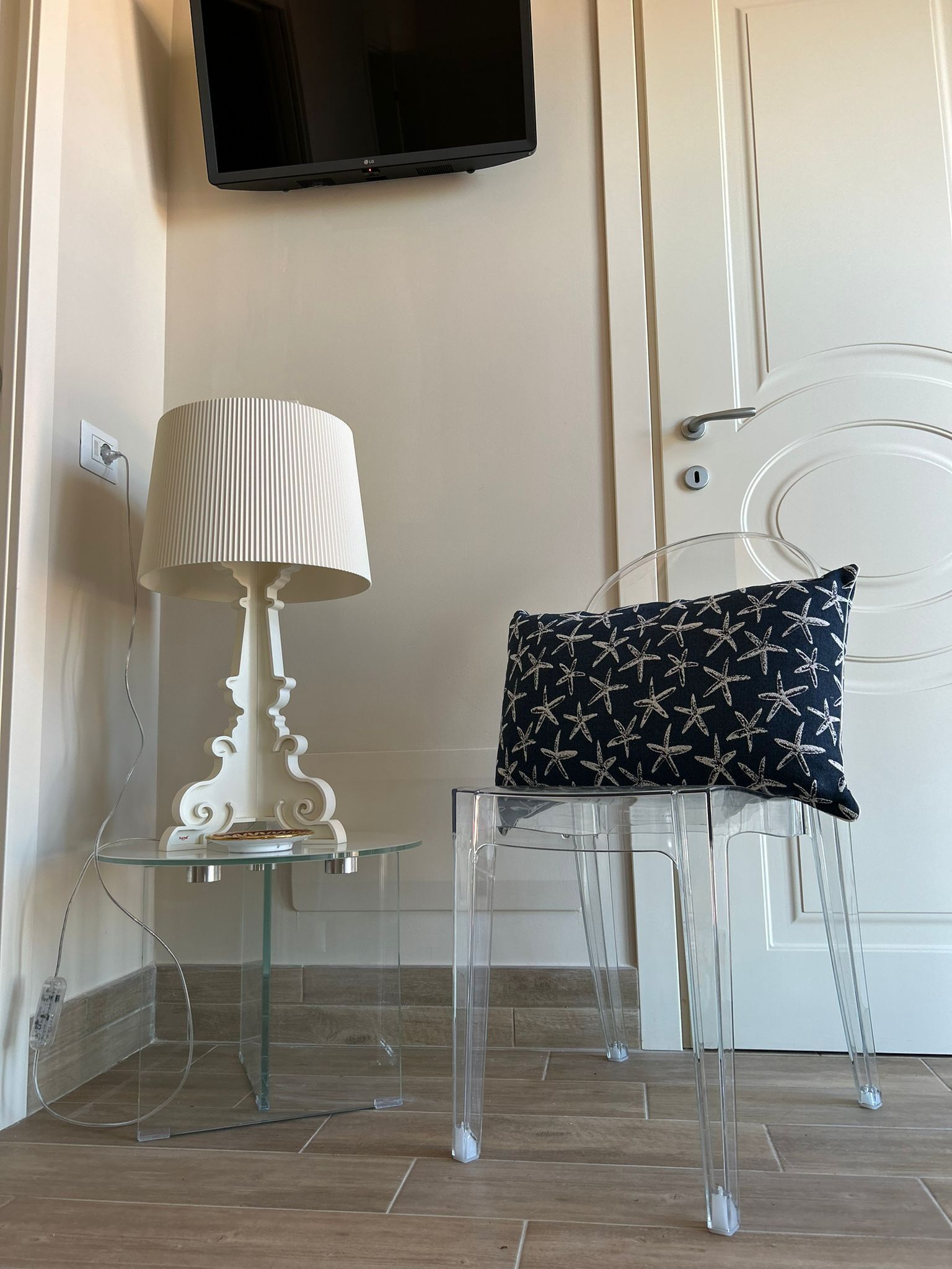 Una sedia trasparente in vetro con un cuscino decorato, una lampada bianca e un televisore a schermo piatto montato a parete.