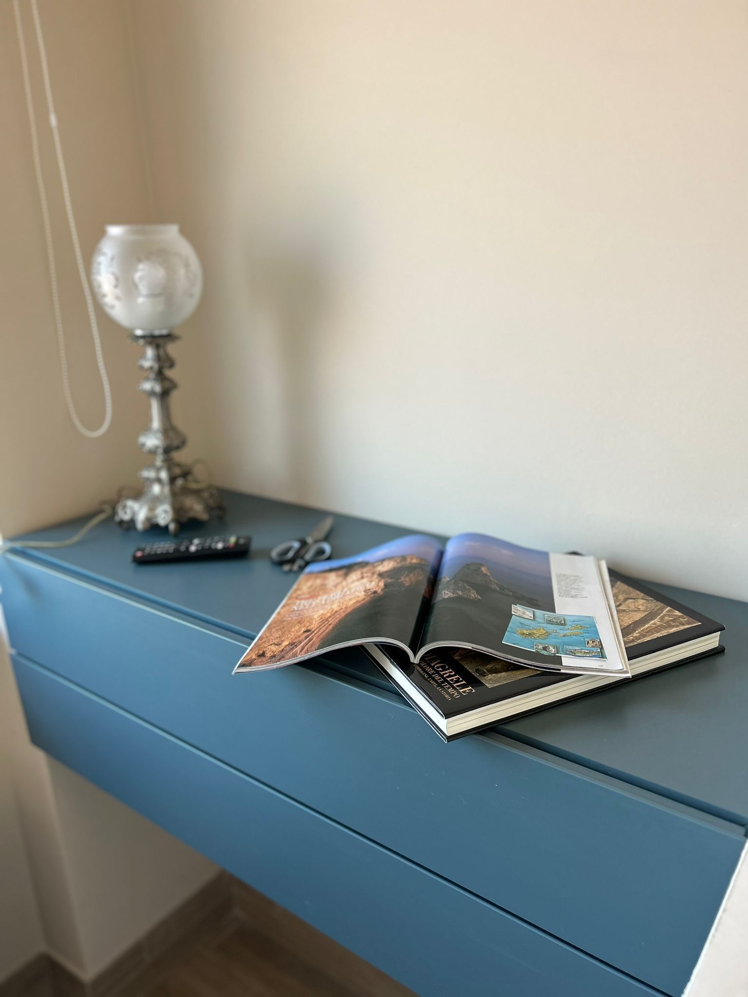 Un escritorio azul con un control remoto, una lámpara de cristal y una revista abierta.