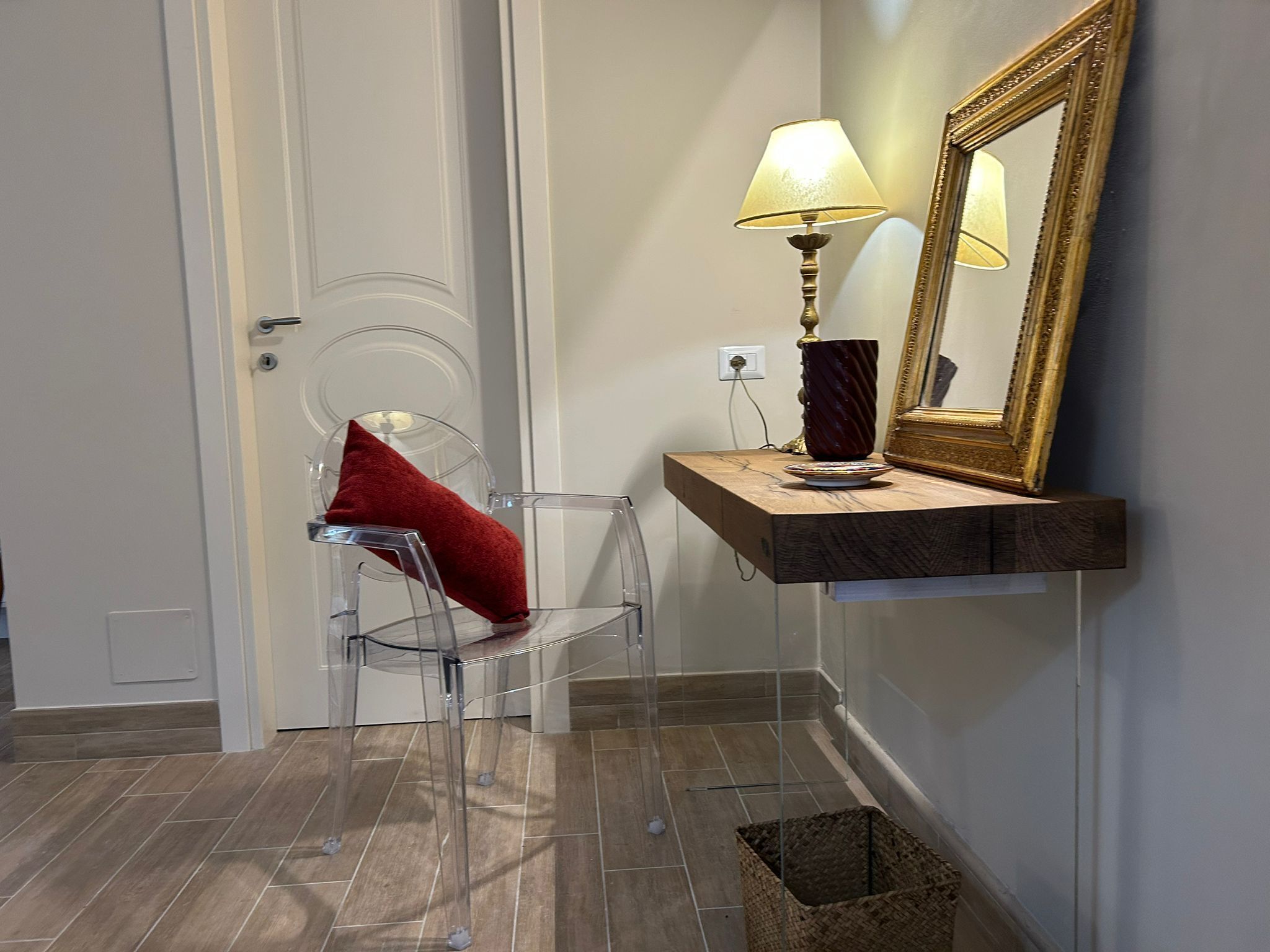 Una habitación con una silla transparente, una lámpara, un espejo y una pequeña cesta.