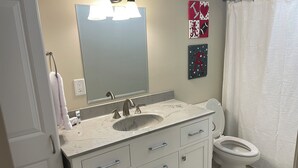 Ein Badezimmer mit Waschbecken, Spiegel und Toilette. Handtücher hängen an der Tür, ein Duschvorhang ist angebracht und ein Schild ist an der Wand befestigt.