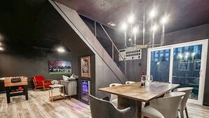 Ein moderner Essbereich mit einem Holztisch, grauen Stühlen und einer Kronleuchter. Im Hintergrund steht ein Tischfußballtisch und ein roter Sessel.