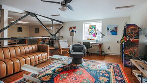 Ein Wohnzimmer mit braunem Ledersofa, einem grauen Sessel, einem farbenfrohen Teppich, einem Deckenventilator und einer TV-Konsole mit einem Poster.