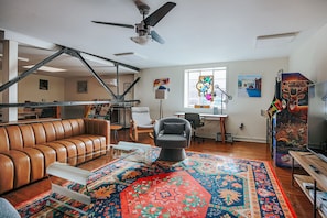Een woonkamer met een bruine leren bank, een grijze fauteuil, een kleurrijk vloerkleed, een plafondventilator en een tv-meubel met een poster.