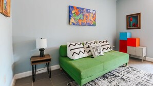 Een moderne slaapkamer met een groene bank, een klein nachtkastje met een lamp, en een kleurrijk abstract schilderij aan de muur.