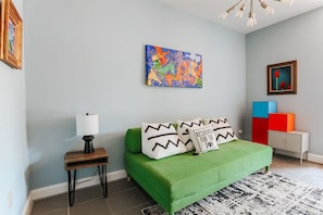 Een moderne slaapkamer met een groene bank, een klein nachtkastje met een lamp, en een kleurrijk abstract schilderij aan de muur.