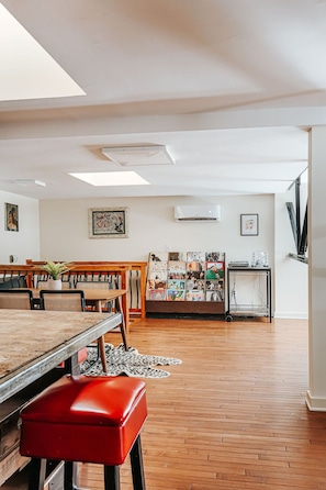 Een ruime kamer met een houten vloer, een eettafel met stoelen, een rode bank, een wandairconditioner en een plank met diverse spullen.