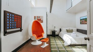 Ein kleines Hotelzimmer mit einem Bett, einer Sitzecke mit einem Sessel, einem kleinen Beistelltisch und einem an der Wand hängenden Kunstwerk.