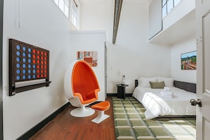 Een compacte hotelkamer met een bed, een zithoek met een stoel, een klein bijzettafeltje en een kunstwerk aan de muur.