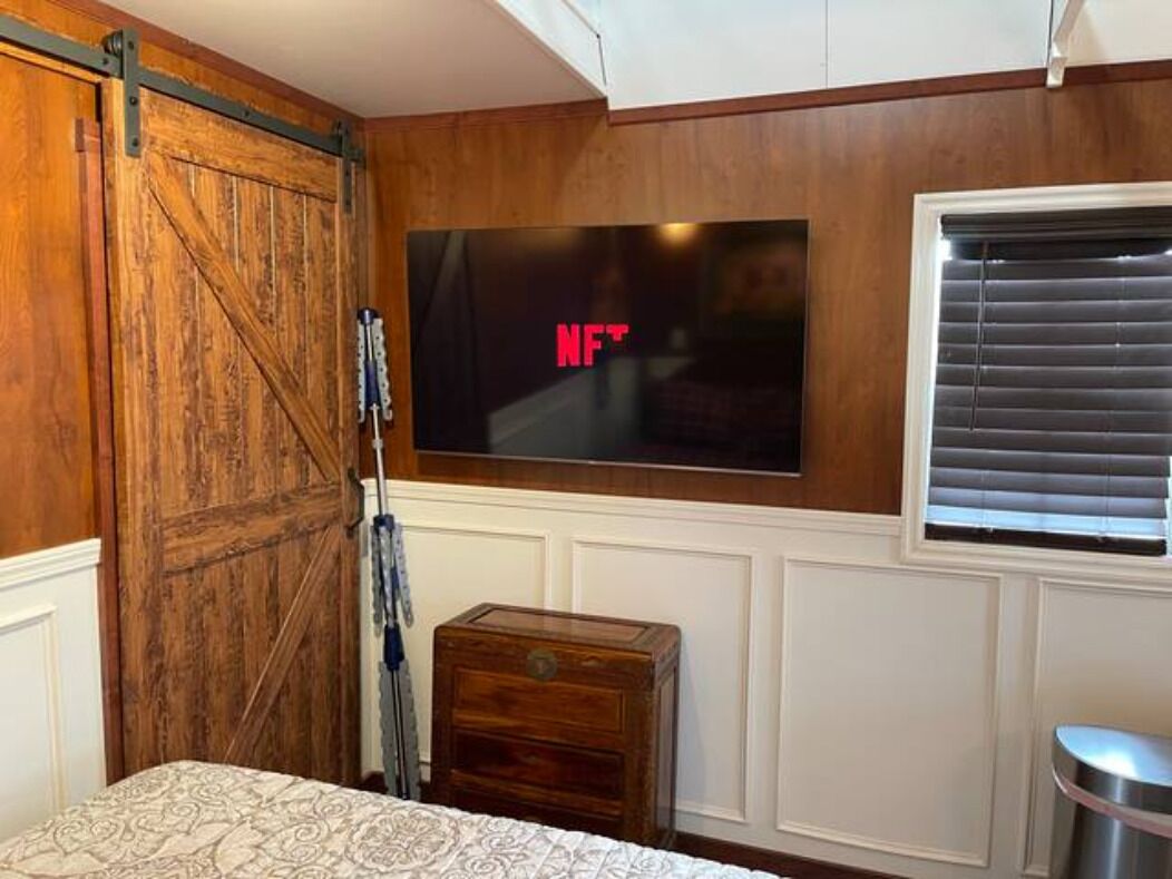 A bedroom with a wooden wardrobe, a TV mounted on the wall, a small wooden chest, and a window with blinds.