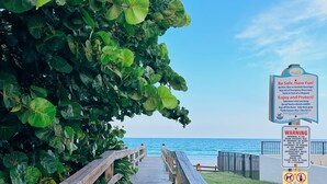 Un paseo marítimo de madera que conduce a una playa, rodeado de exuberante vegetación y cielos despejados.