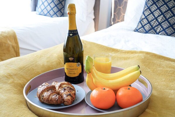 A breakfast tray with croissants, a bottle of Prosecco, bananas, oranges, and a glass of orange juice on a bed with a yellow blanket and patterned pillows.