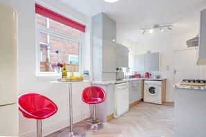A compact kitchen with a washing machine, a sink, and a window with a red blind. There are two red bar stools and a small table with fruit and a vase. - City Centre House • Sleeps 7 • Free Parking • Fast Wifi • Sky TV • Netflix • Self-Check-In • Yoko Property (Northampton)