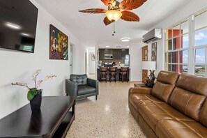 Un salon avec un canapé, un fauteuil, un téléviseur, un ventilateur de plafond, et une cuisine à l’arrière-plan.