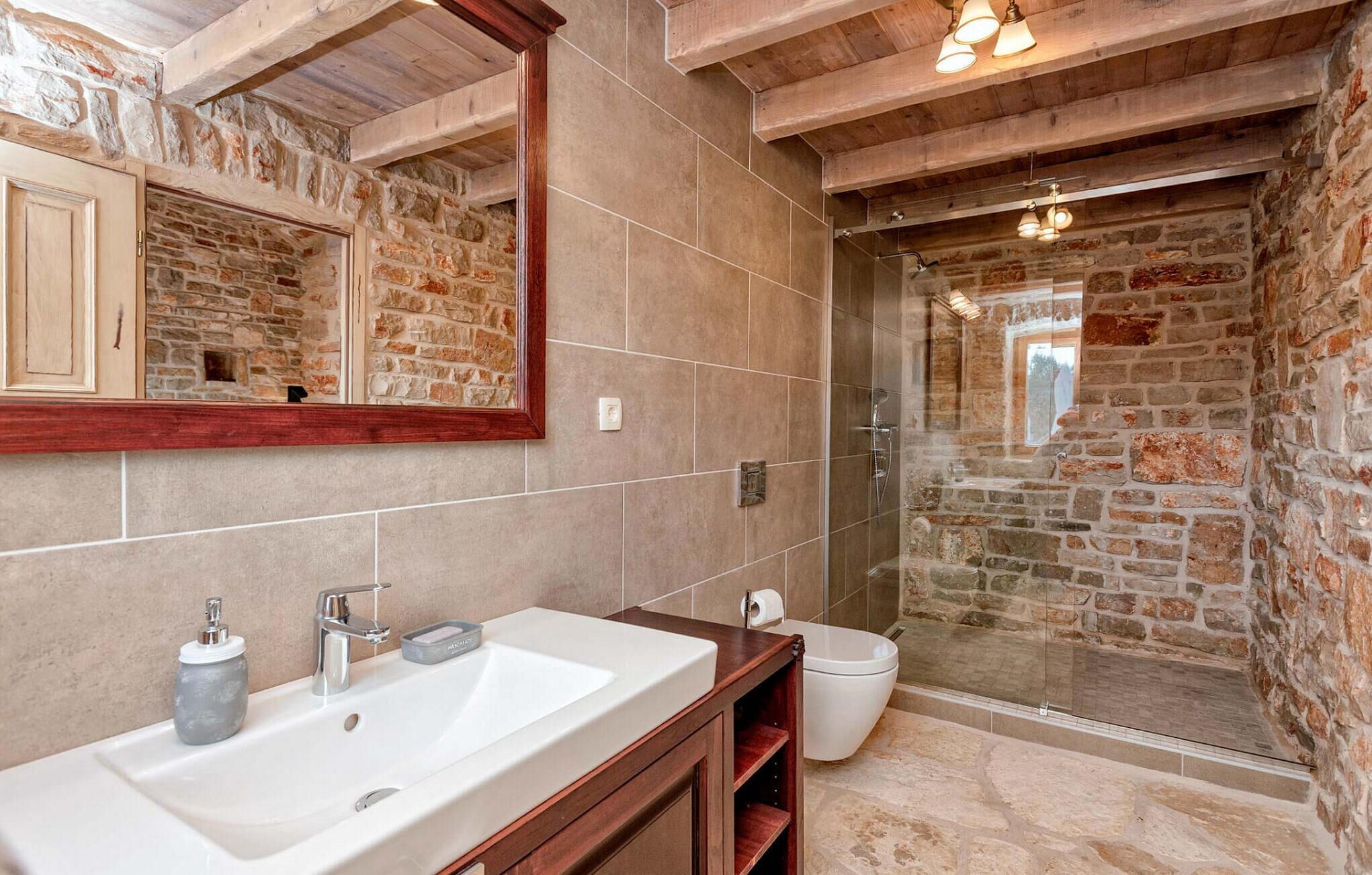 A bathroom with a stone wall, a wooden vanity, a large mirror, and a shower area.
