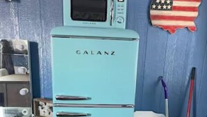 Un refrigerador Galanz color turquesa en una habitación con paredes azules, un armario blanco y una escoba.