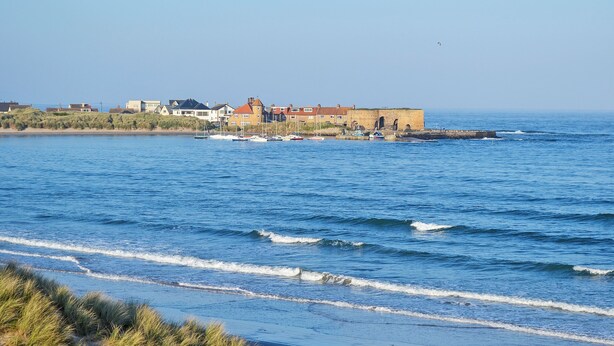Une vue sur la côte, avec une plage de sable, une rangée de maisons et une structure fortifiée au bord de l’eau.