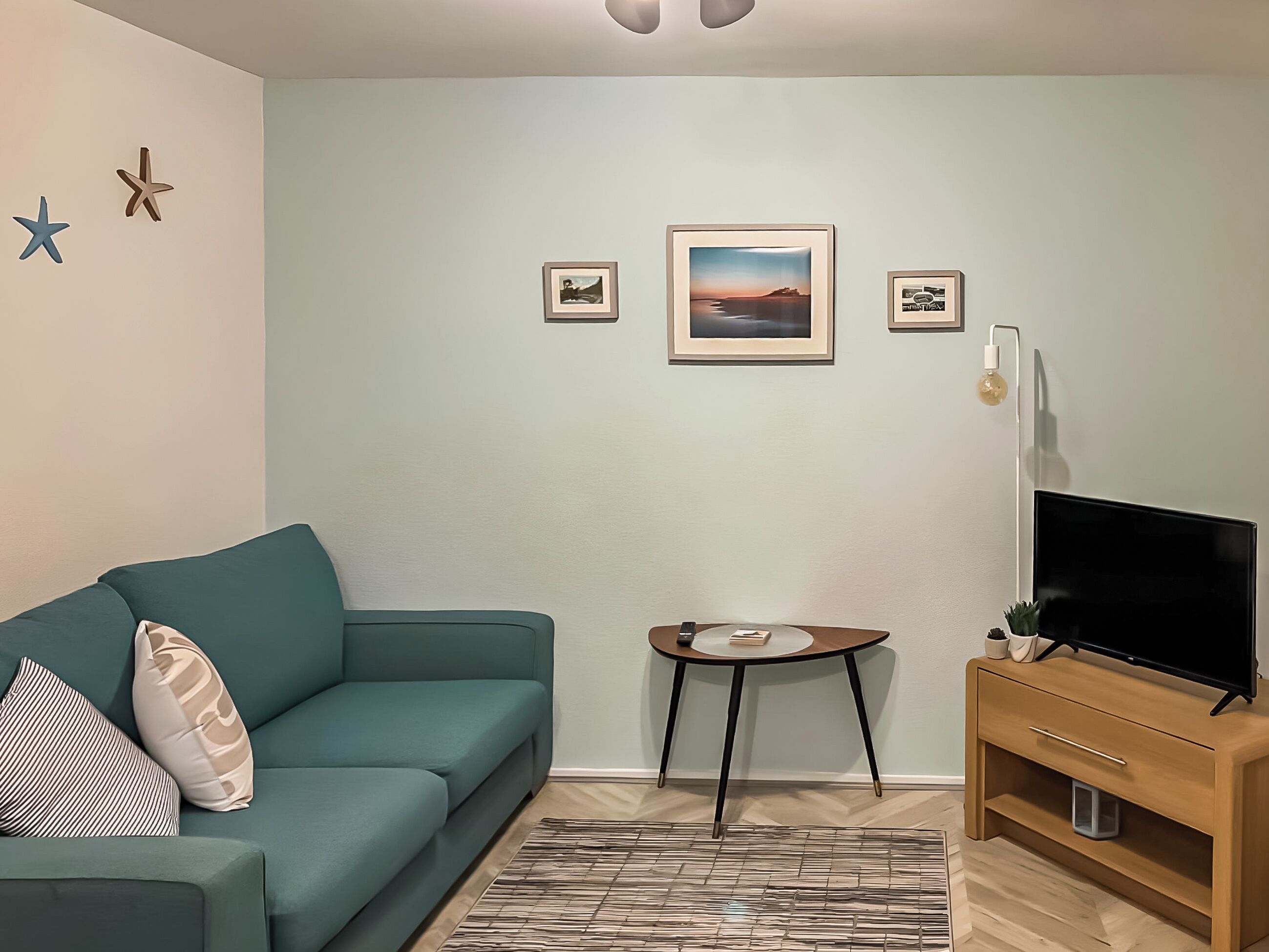A living room with a teal sofa, a small round table, a TV on a wooden stand, and framed pictures on the wall.