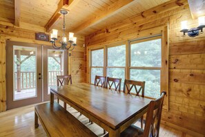 A wooden cabin interior with a large dining table, four chairs, a chandelier, and large windows offering a view of the forest. - Sevierville Rental Cabin w/ Hot Tub & Game Room! (Sevierville)