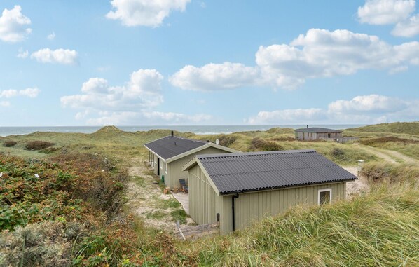 A coastal building with a flat roof, surrounded by dunes and vegetation.
